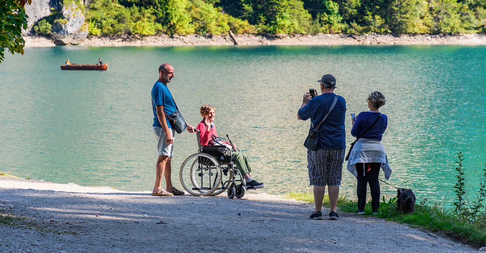 Woman in wheelchair with husband taking a photo in front of a lake.