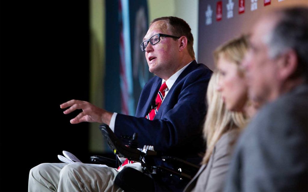 John Morris speaking from a stage while seated in his wheelchair.