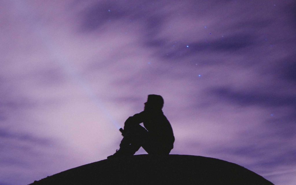 Silhouette of a man sitting on a hilltop in front of a purple night sky.